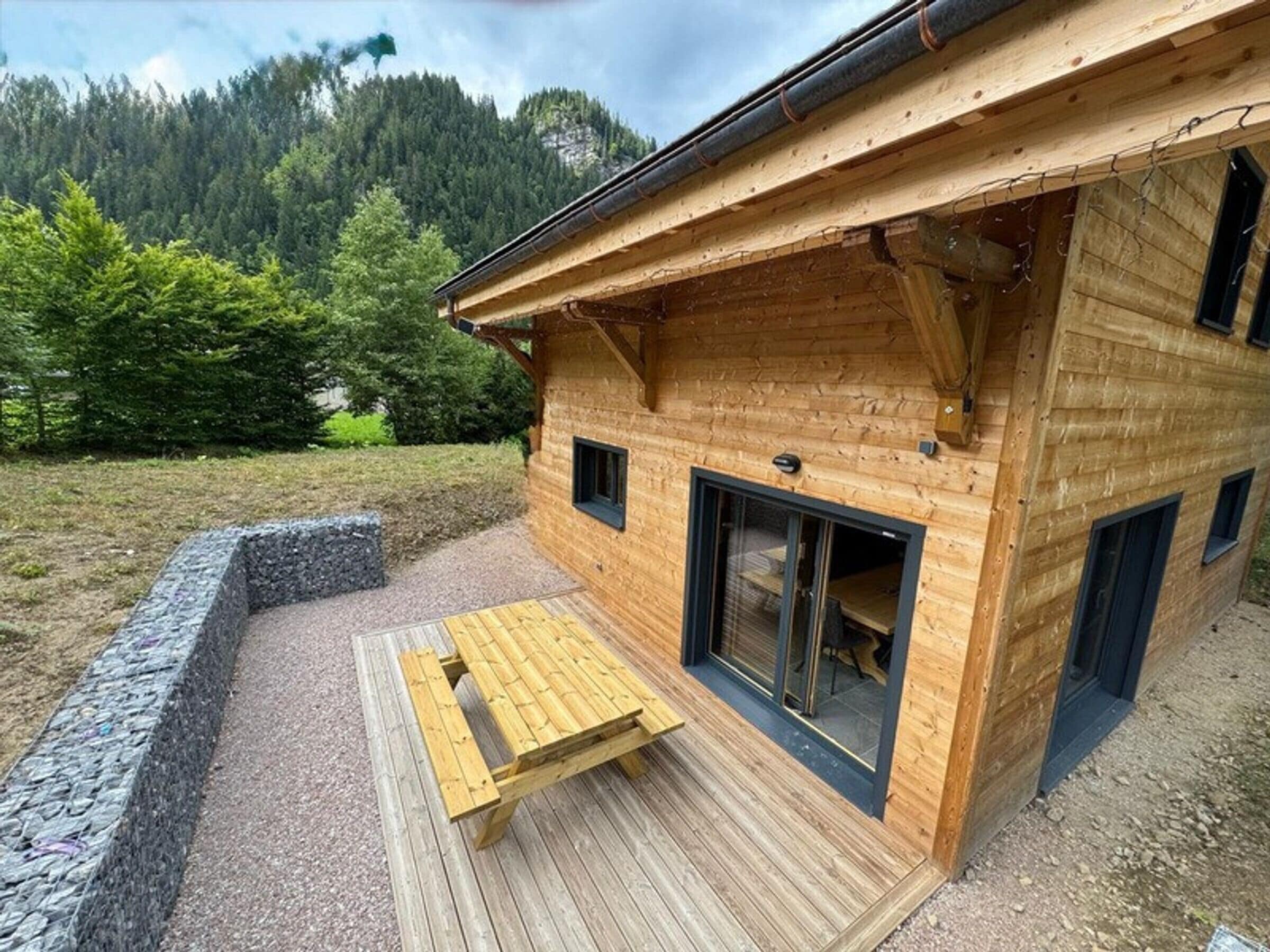 Une cabane en bois dotée d’une terrasse, d’un mur en pierre et offrant une vue sur une montagne boisée.