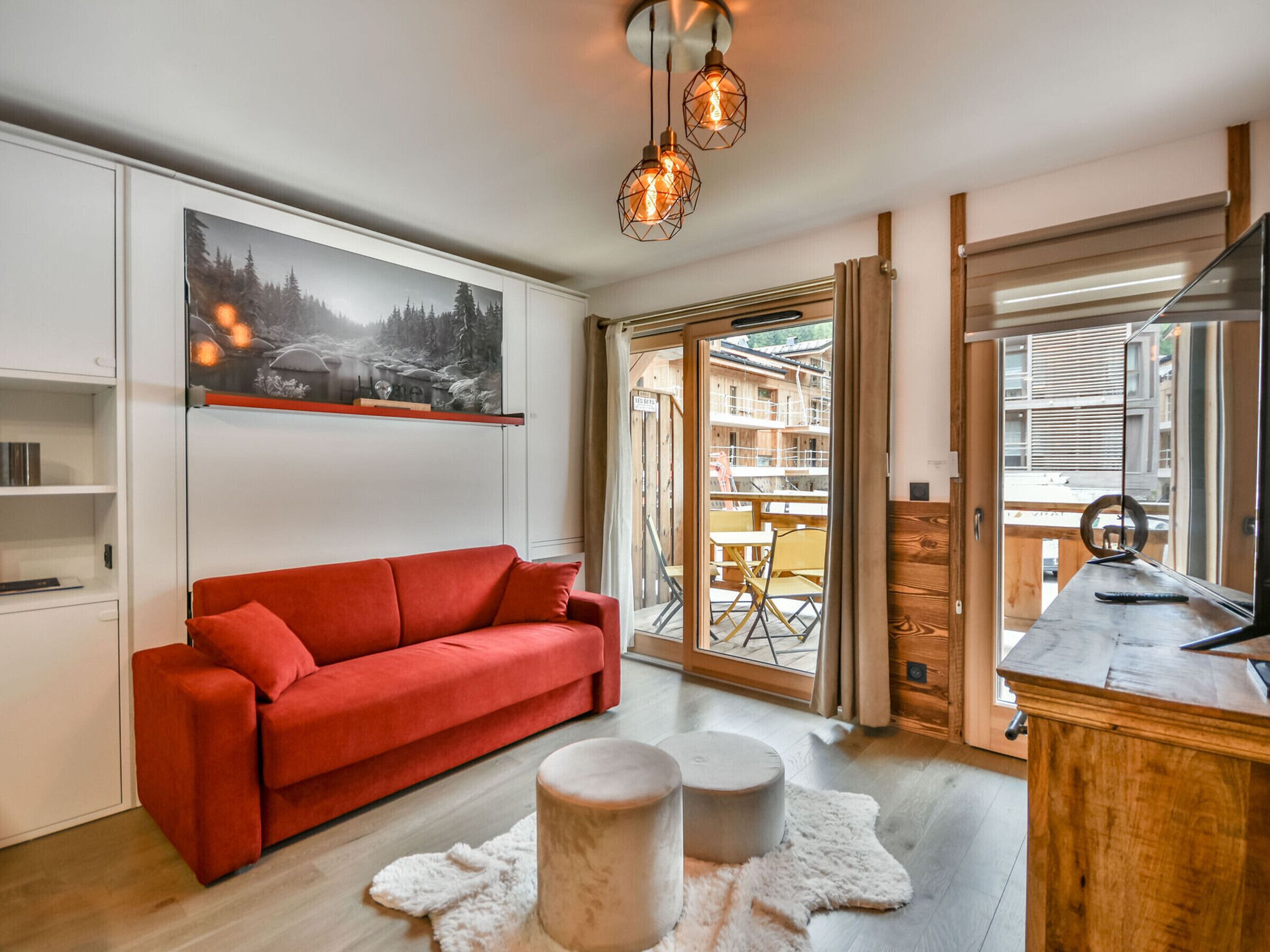 Een moderne woonkamer met een rode bank, houten vloer, een groot raam met uitzicht op een gebouw en een schilderij aan de muur.