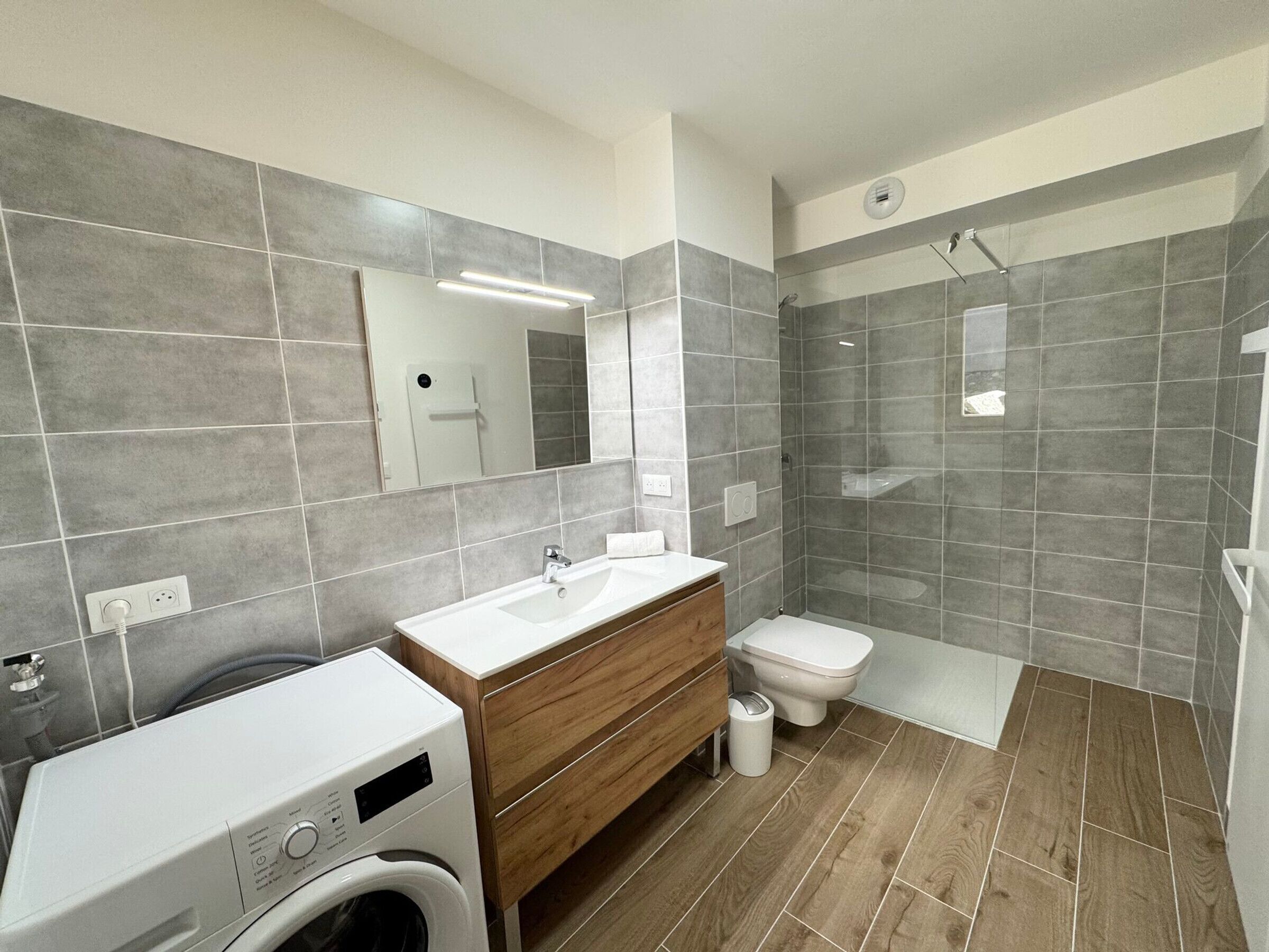 Une salle de bain moderne équipée d’un lave-linge intégré, d’un meuble-lavabo en bois, de toilettes et d’une cabine de douche en verre.