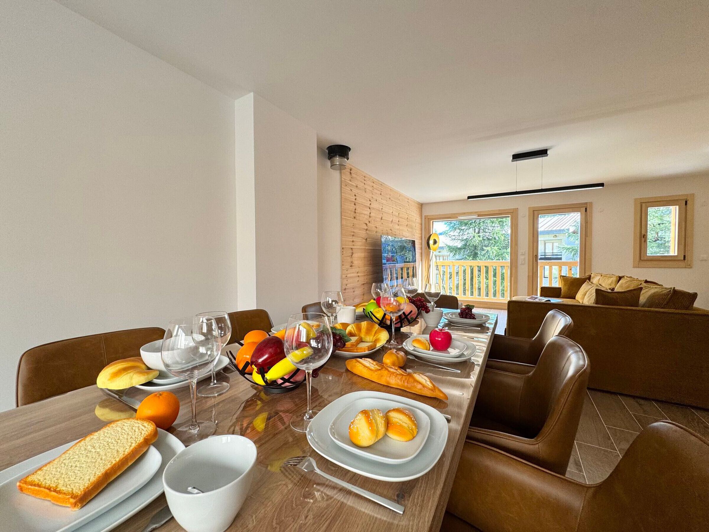 A dining area with a long table set for a meal, including bread, fruits, and wine glasses.