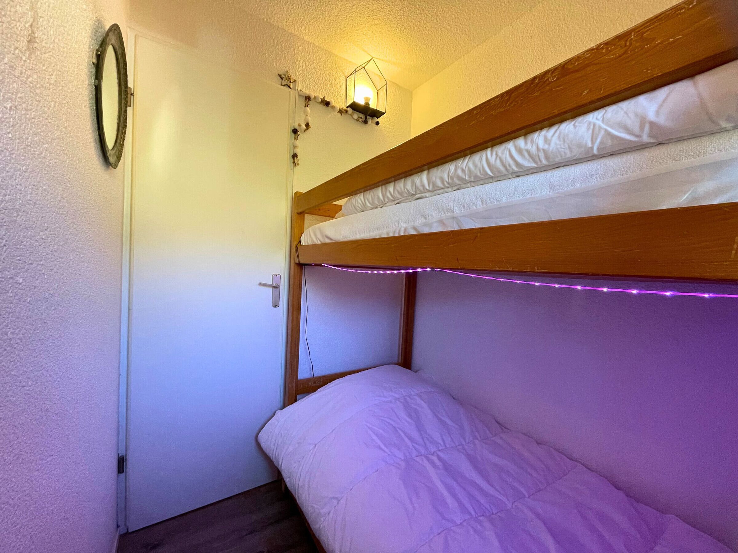 A bunk bed with a wooden frame and white bedding. There is a wall-mounted light fixture above the bed and a door to the left.