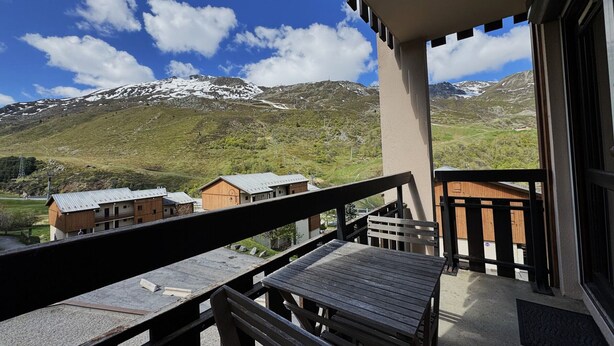 Ein Balkon mit Holztisch und -stühlen, der auf eine gebirgige Landschaft mit schneebedeckten Gipfeln und blauem Himmel blickt.