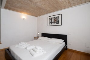 A neatly made bed with white linens and towels, a black headboard, a framed picture on the wall, and a small bedside table with a lamp.