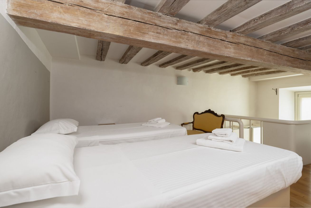 A room with two beds, wooden beams on the ceiling, a chair, and a window.