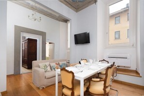 Un comedor con una mesa blanca preparada para cuatro personas, rodeada de sillas de madera con asientos acolchados. Un televisor está montado en la pared sobre la mesa.