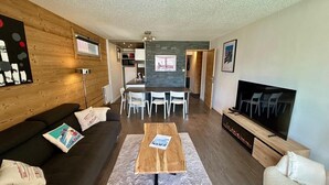 A modern living room with a dark sofa, a wooden coffee table, and a TV. There is a dining area with a table and chairs, and a kitchen in the background. - Appartement-pied des pistes -T3 -60 m2 -Garage _ Free WFI - Resort center (Les Deux Alpes)