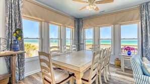 A dining area with a table and chairs, large windows with a view of the beach, and a ceiling fan.