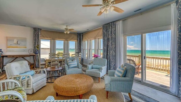 A spacious living room with a large window overlooking a beach, furnished with striped chairs and a round wicker ottoman.