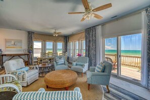 A spacious living room with a large window overlooking a beach, furnished with striped chairs and a round wicker ottoman. - Lemontiniville  - Oceanfront - Decks for Days- Dog Friendly (Holly Ridge)