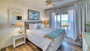 A bedroom with a bed, a ceiling fan, a nightstand, a lamp, a painting, and a view of the ocean.