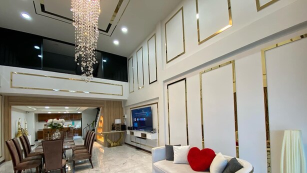 A spacious living area with a high ceiling, a chandelier, a sofa with a heart-shaped pillow, a TV, and a dining area with chairs and a table. - Peema Villa (U Thong)