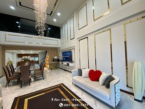 A spacious living area with a high ceiling, a chandelier, a sofa with a heart-shaped pillow, a TV, and a dining area with chairs and a table. - Peema Villa (U Thong)