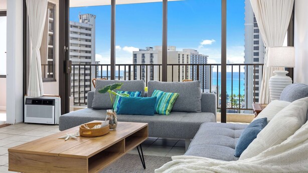 A modern living room with a grey sofa, a wooden coffee table, and a view of the ocean through large windows.