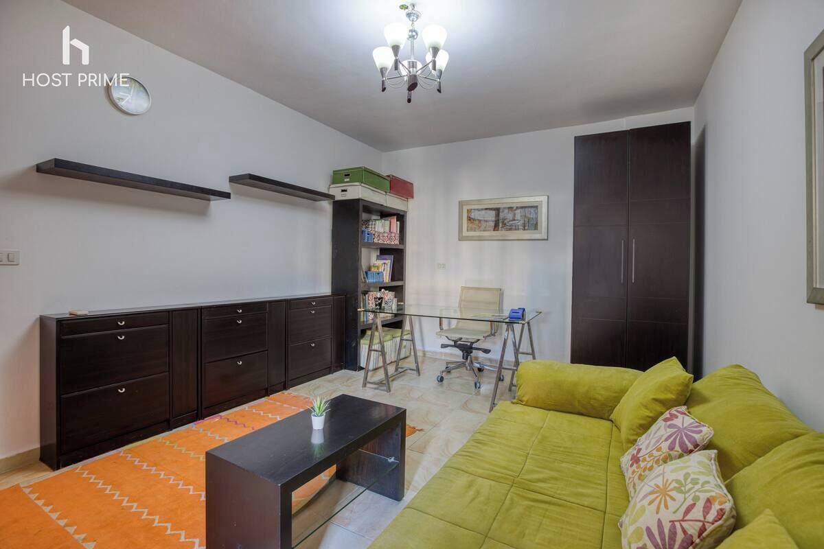 A modern living room with a green sofa, a coffee table, a bookshelf, a desk with a chair, and a dark wardrobe.