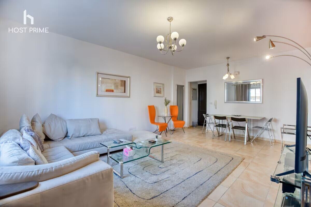 A modern living room with a sofa, glass coffee table, and orange chairs. There is a dining area with a table and chairs.