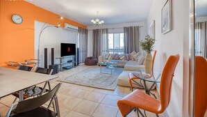 A modern living room with a sofa set, a dining table with chairs, a flat-screen TV, and a large window with curtains. - Comfy Apartment in Rehab City (New Cairo)