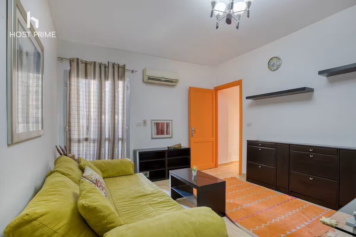 Een moderne woonkamer met een groene bank, een klein tafeltje en een vloerkleed met een patroon. Er hangt een airconditioner aan de muur en een deur leidt naar een andere kamer.