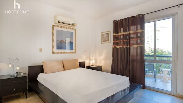 A bedroom with a bed, bedside tables, an air conditioning unit, a window with curtains, and a view of the outdoors. - Comfy Apartment in Rehab City (New Cairo)