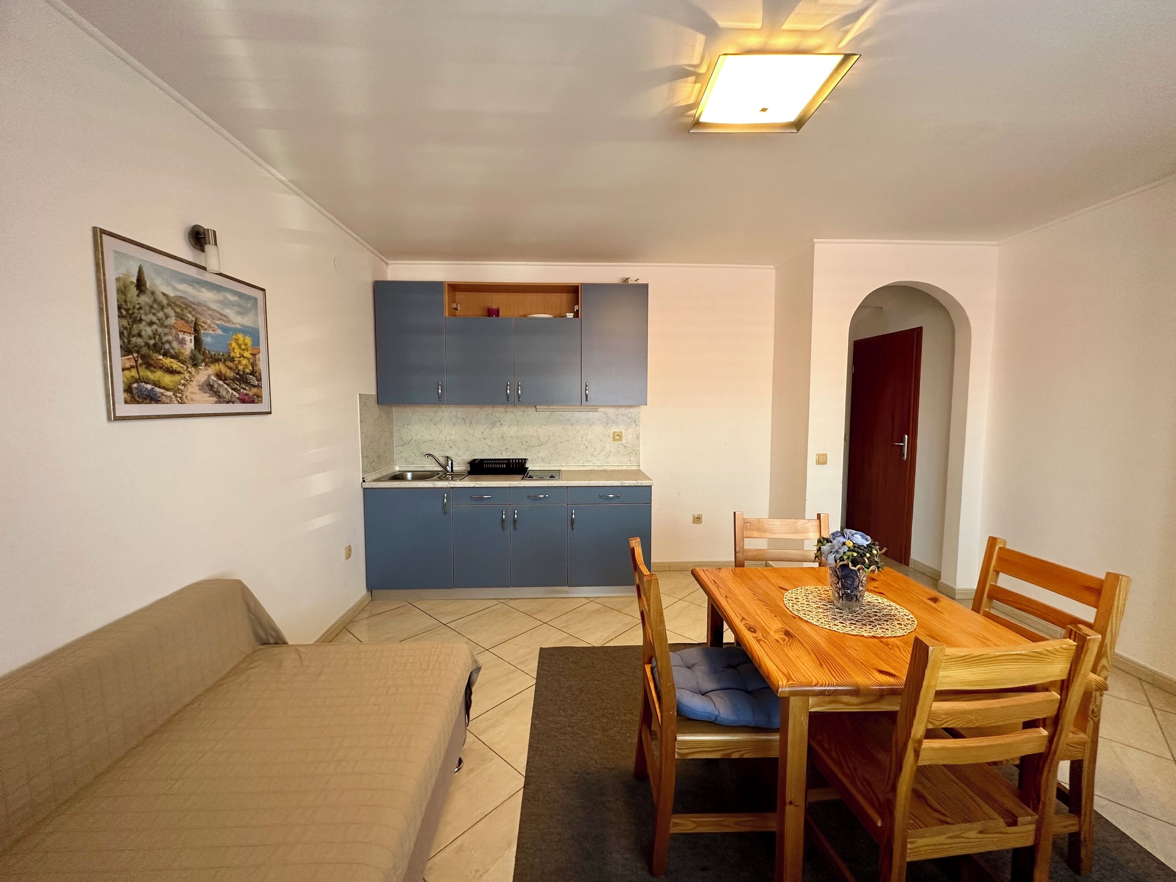 A compact kitchen with blue cabinets, a dining table with wooden chairs, and a painting on the wall.