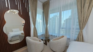 Une chambre d’hôtel moderne, dotée d’un grand miroir, d’un lit, de deux fauteuils, d’une petite table ronde et de grandes fenêtres avec des rideaux légers.