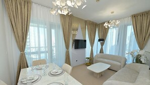 Un salon moderne comprenant un coin repas, des rideaux beiges, un téléviseur à écran plat et un lustre.