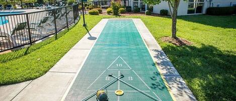 A shuffleboard court with a green surface, a pool table, and a fenced area with lounge chairs.