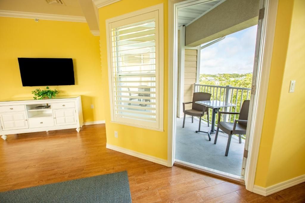 A room with a wooden floor, a white entertainment unit, a flat-screen TV, a sliding glass door leading to an outdoor balcony with a table and chairs.