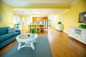 A modern living room with a blue sofa, a glass coffee table, and a kitchen area with wooden cabinets and a refrigerator.