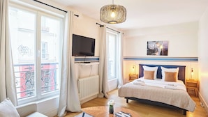 Une chambre d’hôtel comprenant un lit, une télévision, une table de chevet, une chaise, une fenêtre avec des rideaux et un luminaire suspendu.