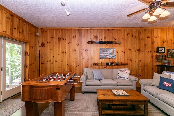 A cozy living room with wooden walls, a foosball table, a sofa with family-themed pillows, and a coffee table.