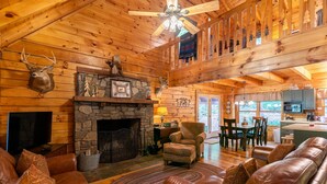 A cozy cabin interior with a stone fireplace, leather sofas, a dining area, and a kitchen visible through a glass door. - Garretts Getaway at Watauga Lake - Private Dock, Lakefront (Butler)