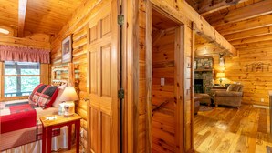 A cozy log cabin interior with a wooden bed, a small table with a lamp, and a fireplace in the background. - Garretts Getaway at Watauga Lake - Private Dock, Lakefront (Butler)