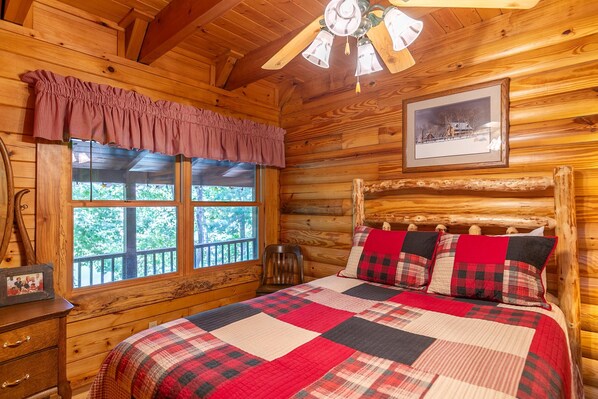 A wooden cabin room with a bed, a ceiling fan, a window with a view of trees, and a framed picture on the wall.