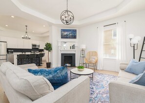Un salon moderne avec un canapé, une table basse et une cheminée. On aperçoit au fond une cuisine avec des meubles blancs et un coin repas.