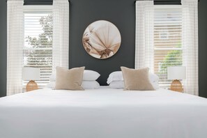 A bedroom with a large bed, two bedside tables with lamps, a round wall mirror, and white curtains.