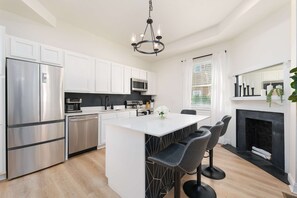 A modern kitchen with stainless steel appliances, a central island with bar stools, and a fireplace. - The Laura | Entire Historic Home | Main St (Jacksonville)