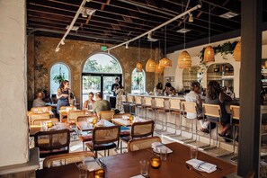 A restaurant interior with high ceilings, exposed beams, and natural light from large windows. Tables are set with candles and glasses, and patrons are dining.