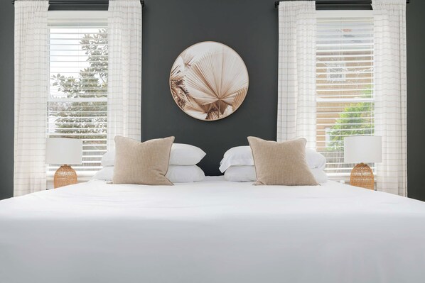 A bedroom with a large bed, two bedside tables with lamps, a round wall mirror, and white curtains.