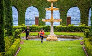 Een tuin met een fontein, hagen en een waterpartij in de verte.