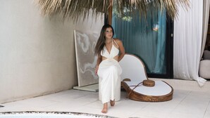 A woman sitting by a poolside with a thatched roof overhead, a white chair, and a wooden stool.