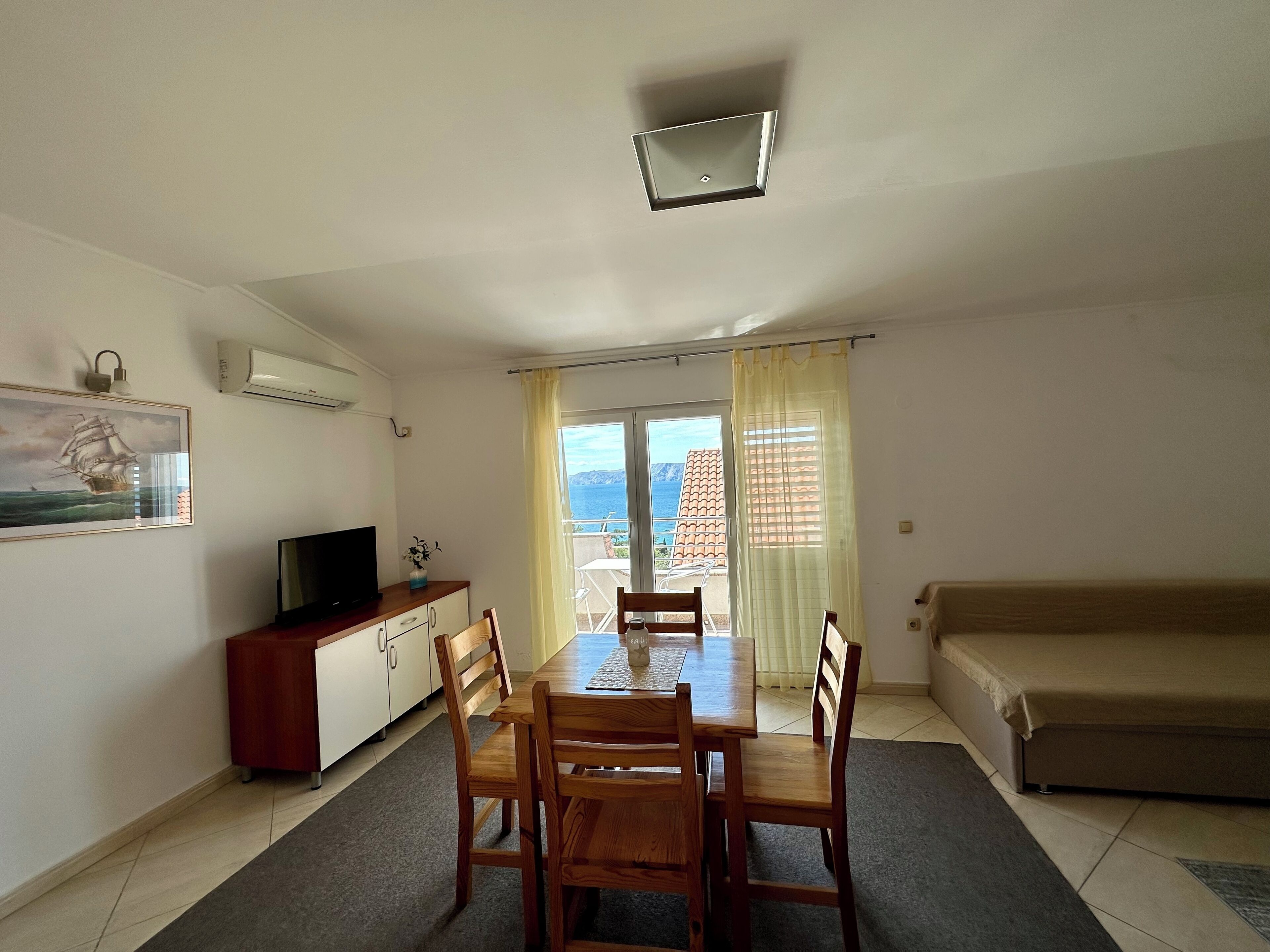 A living room with a dining table, chairs, a sofa, a TV on a cabinet, and a view of the sea through a glass door.