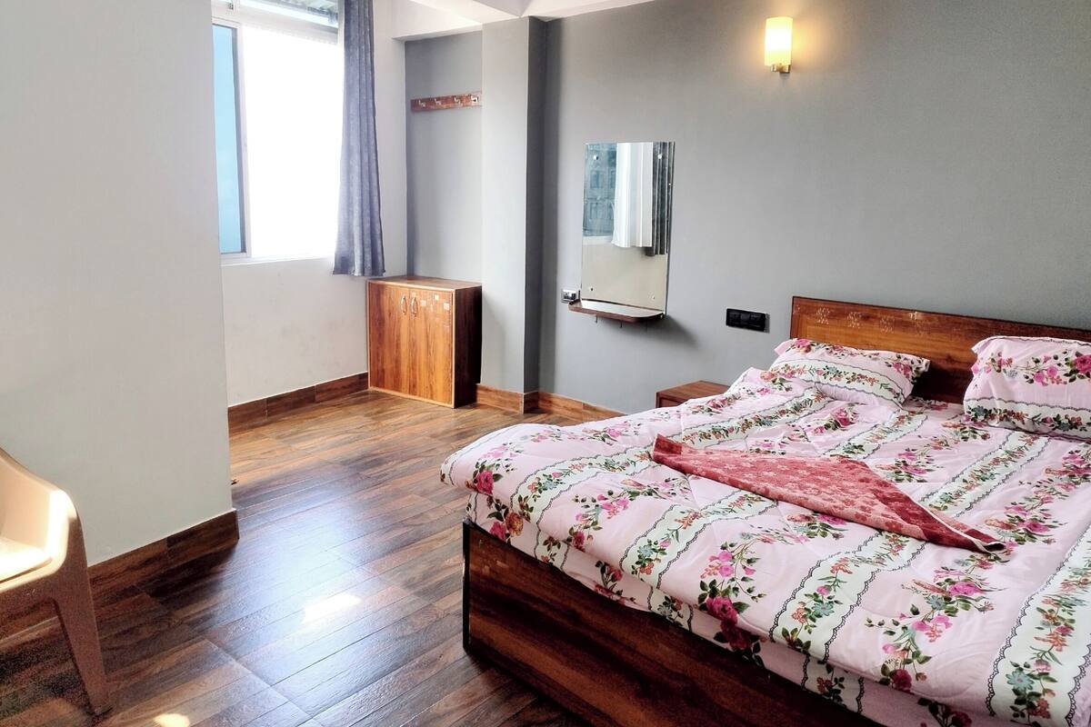 A bedroom with a wooden bed, a small wooden cabinet, a mirror, and a window with curtains.