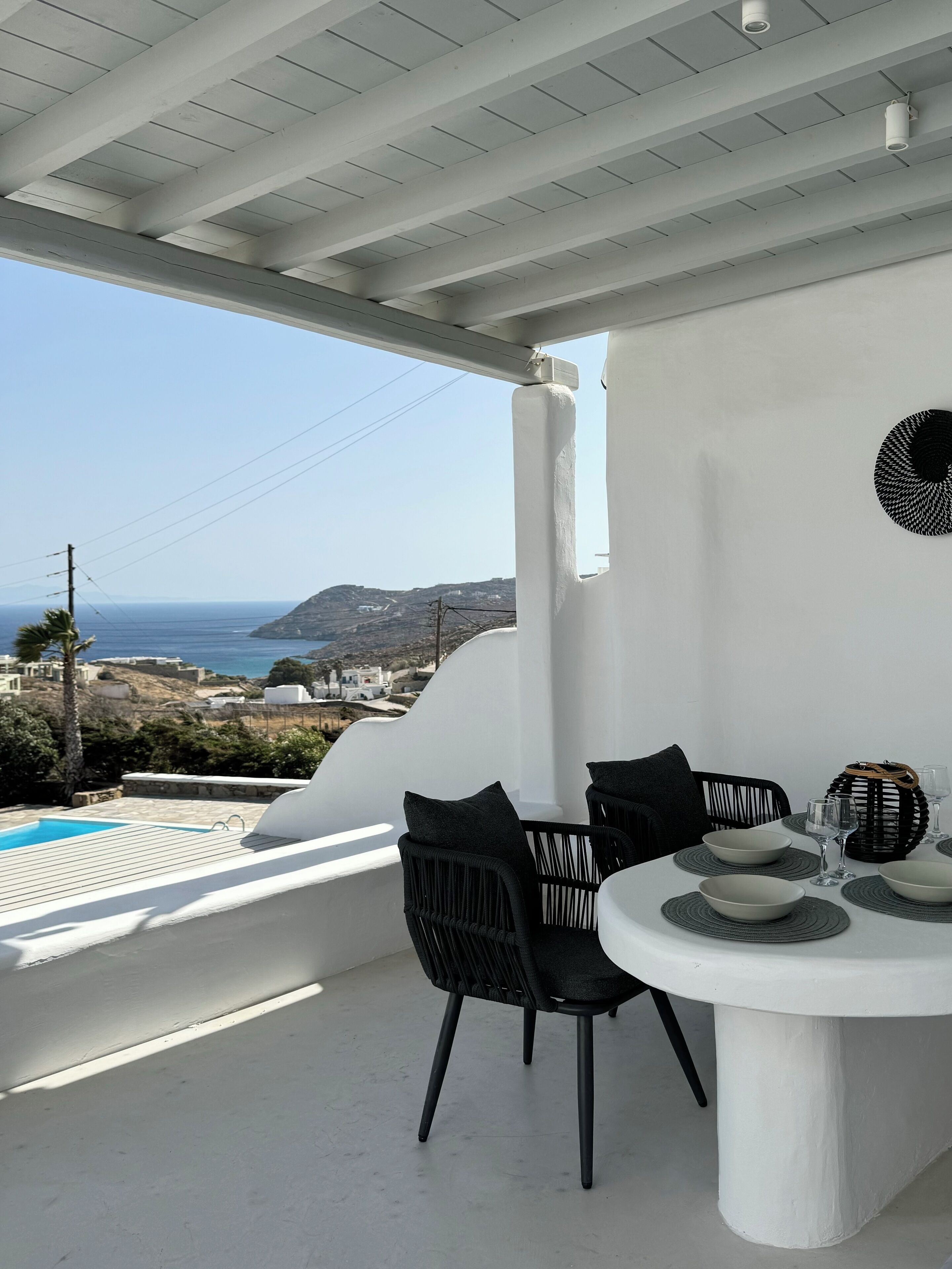 A balcony with a white table set for dining, black chairs, and a view of the sea and hills.