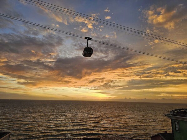 Eine Seilbahn fährt über ein Gewässer, im Hintergrund ist ein Sonnenuntergang zu sehen.