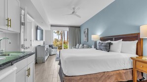 Una habitación de hotel moderna con una cama grande, cabecera de madera, mesita de noche, escritorio, silla, un amplio ventanal con cortinas y una zona de cocina con armarios blancos y un lavabo.