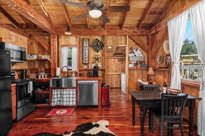 A rustic cabin interior with a kitchen, dining area, and a large window. - Texas Saw Mill (Cabin #1) - Hot Tub & 10 Mins to Main St (Fredericksburg)
