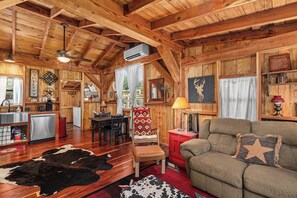 A rustic cabin interior with wooden beams, a dining area, a kitchen, and a living room with a sofa and a cowhide rug. - Texas Saw Mill (Cabin #1) - Hot Tub & 10 Mins to Main St (Fredericksburg)