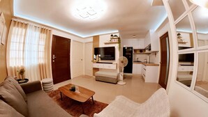 Una sala de estar moderna con un sofá, una mesa de centro y un televisor. Hay una zona de cocina con un microondas y un refrigerador.