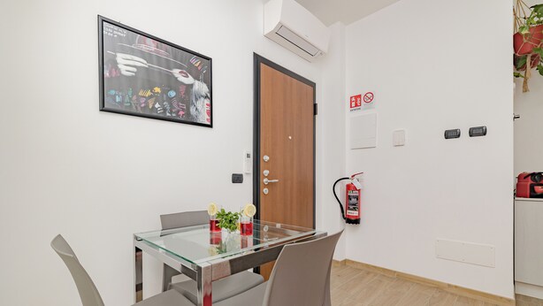 A modern dining area with a glass table, grey chairs, a framed artwork, a wooden door, and a fire extinguisher on the wall.