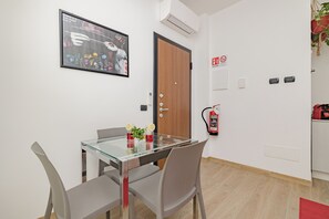 A modern dining area with a glass table, grey chairs, a framed artwork, a wooden door, and a fire extinguisher on the wall.